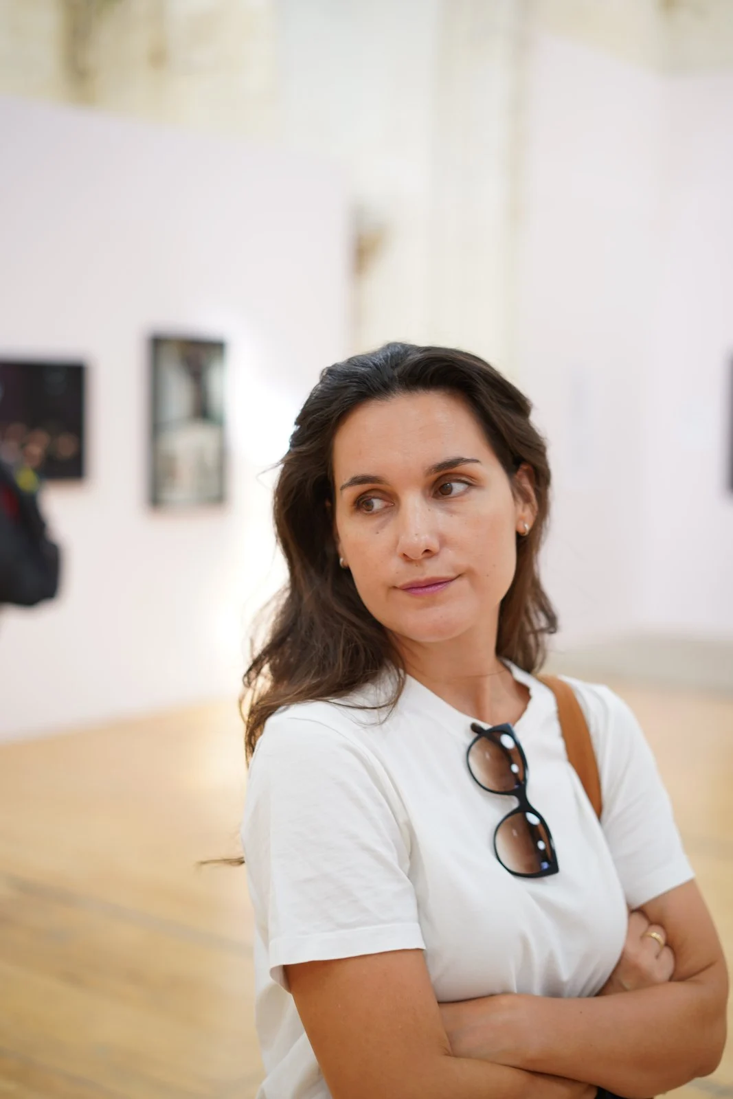 Kvinna i vit t-shirt med solglasögon i håret står i ett ljust gallerirum med vita väggar