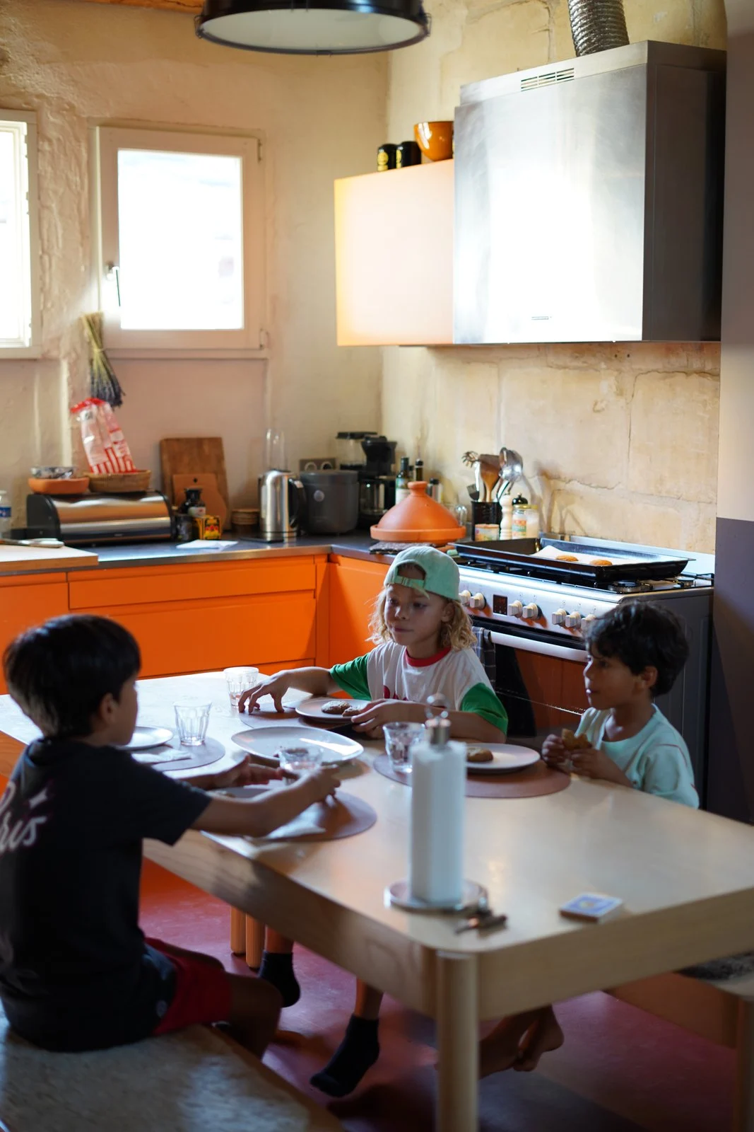 Tre barn vid matbord i ett kök med orange skåp, vit spis och gula väggar